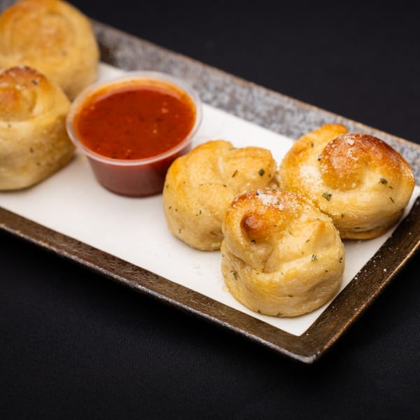 Golden-brown garlic knots with herbs and Parmesan cheese, served with a side of marinara sauce.