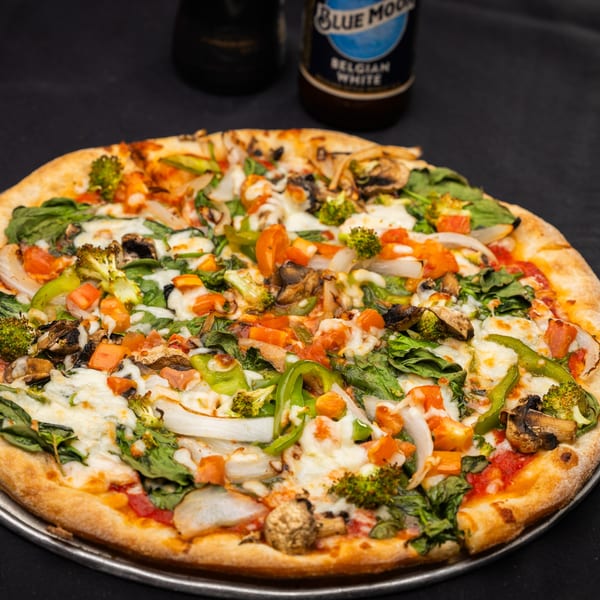 A vibrant I Love NY Pizza veggie pizza with broccoli, spinach, peppers, onions, and mushrooms, with beer bottles in the background.